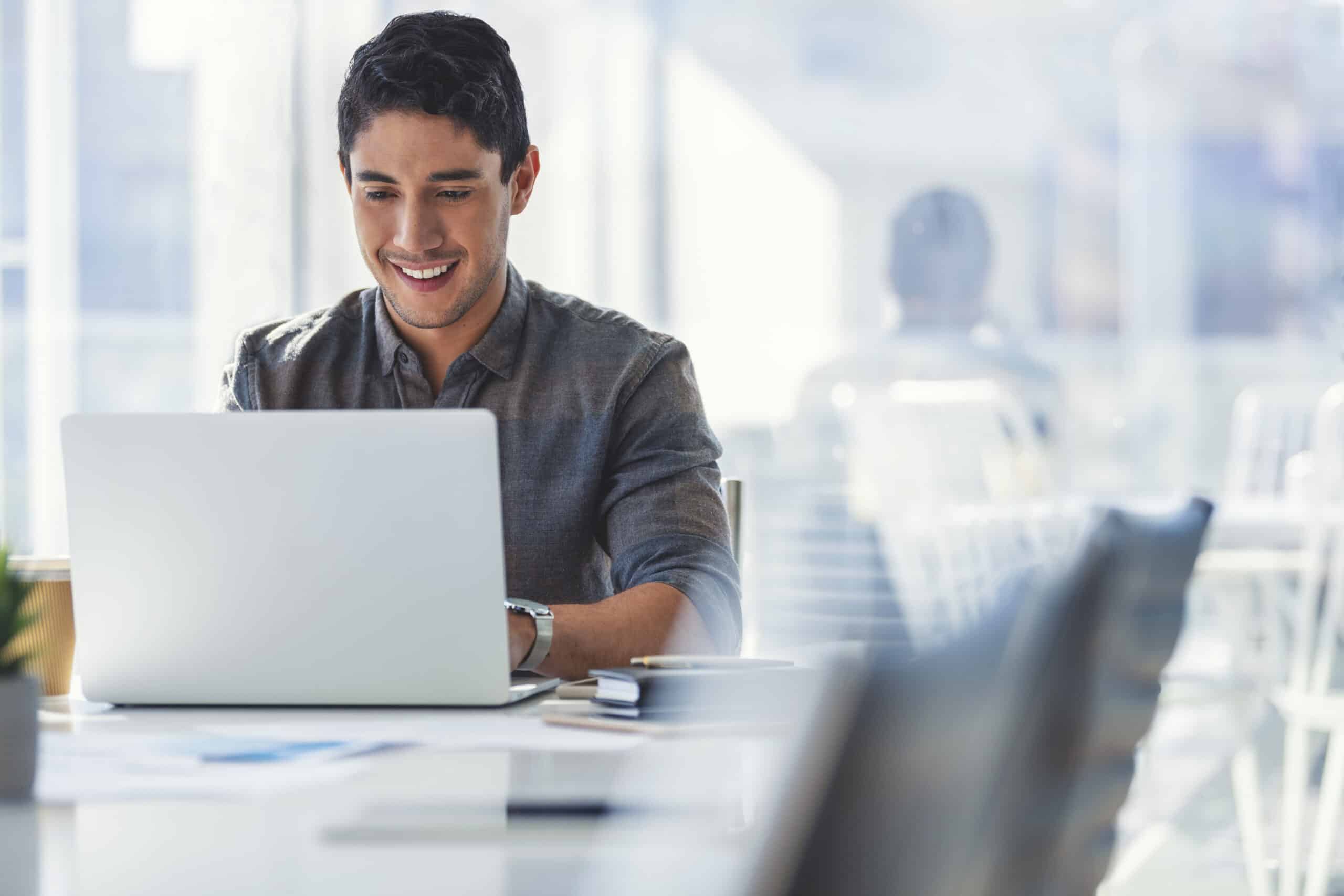 Businessman working on a laptop computer in the office. He is happy and smiling and there is a window behind him. He is casually dressed